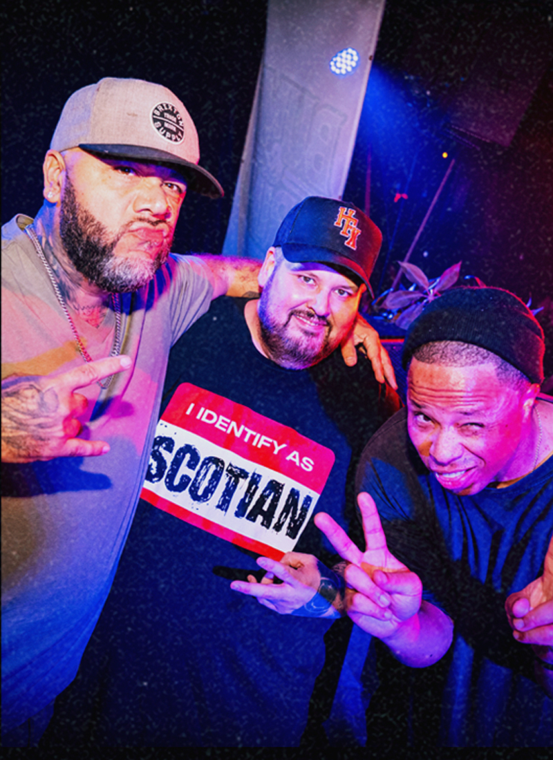 Three friends posing indoors under blue and purple club lighting, one wearing an "I Identify As Scotian" shirt