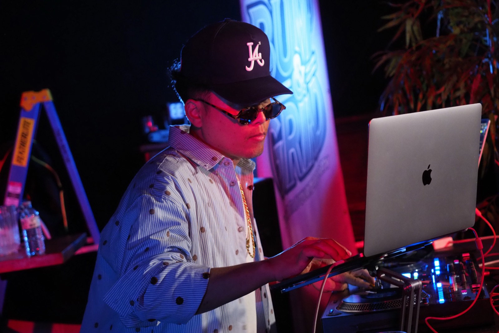A DJ in sunglasses and a cap mixing on a laptop in front of the Bump & Grind banner
